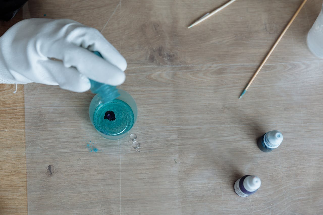 services-04 A gloved hand pouring blue resin into a container on a wooden table, creating art.